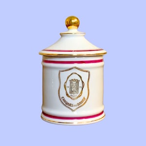 May include: A white ceramic jar with a gold finial and trim. The jar features a shield emblem with the text "CHIQUES de BAVAY" and the year "1875". A red band encircles the jar.