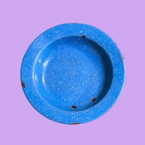Könnte beinhalten: Eine runde, blaue Emaille-Schüssel mit einem weißen Sprenkelmuster auf der Oberfläche. Die Schüssel hat einen breiten, flachen Rand und weist sichtbare Gebrauchsspuren mit kleinen braunen Roststellen an den Rändern und am Boden auf.