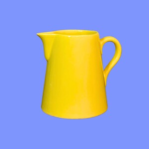 Op de afbeelding: Een felgele keramische kan met een gladde, glanzende afwerking. De kan heeft een brede, taps toelopende vorm, een gebogen handvat aan de rechterkant en een duidelijke schenktuit aan de linkerkant. Het ontwerp is eenvoudig, functioneel en effen.