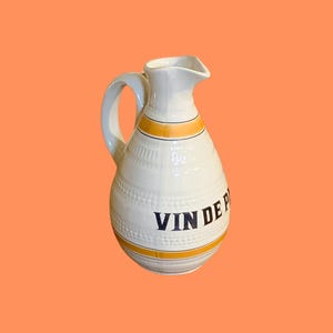 Op de afbeelding: Witte keramische kan met een handvat en tuit, versierd met gele en zwarte strepen. De woorden "VIN DE P" zijn in donkerblauw gedrukt. De kan heeft een gestructureerd oppervlak en een afgeronde vorm.