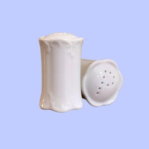 Puede incluir: Dos saleros y pimenteros de cerámica blanca. Los saleros y pimenteros tienen un diseño acanalado en la base y un patrón floral cerca de la parte superior. Uno está en posición vertical y el otro está de lado, revelando los agujeros.