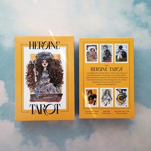 May include: The Heroine Tarot deck box features a yellow background with black text. The front of the box shows an illustration of a woman with long dark hair wearing a white dress and holding a glass of liquid. The back of the box shows a description of the deck and the illustrations.