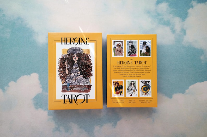 May include: The Heroine Tarot deck box features a yellow background with black text. The front of the box shows an illustration of a woman with long dark hair wearing a white dress and holding a glass of liquid. The back of the box shows a description of the deck and the illustrations.