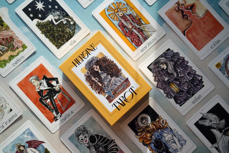 May include: A tarot deck with a yellow box featuring the text "Heroine Tarot". The cards feature illustrations of people and symbols in a dark, whimsical style.