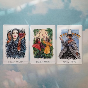 May include: Three tarot cards with watercolor illustrations. The first card is the Three of Swords, depicting a woman in a blue feathered coat with three swords piercing a heart. The second card is the Four of Wands, showing two women in a forest setting, one in a red dress and the other in a yellow dress. The third card is the Seven of Cups, featuring a woman in a black and white dress with a white hat, holding seven cups.