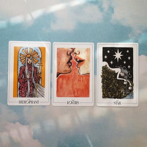 May include: Three tarot cards, The Hierophant, The Lovers, and The Star. The Hierophant card features a woman in a red and white robe holding a staff. The Lovers card shows a couple kissing. The Star card depicts a woman pouring water from two jugs into a pool of water.