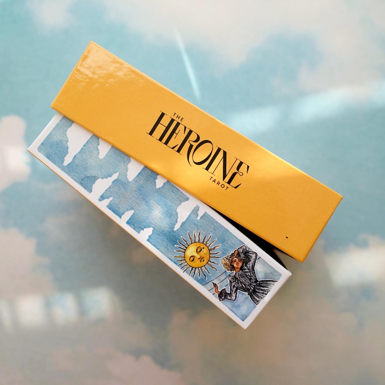 May include: A yellow box with the text "THE HEROINE TAROT" on the top. The box is open and a tarot card with a blue and white watercolor background is visible. The card features a woman in a white dress standing on a cloud with a sun behind her.