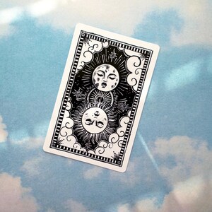 May include: Black and white illustration of a sun and moon with faces, surrounded by clouds and stars. The image is on a white card with a black border.