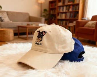May include: A beige baseball cap with an embroidered Schnauzer dog design and the name "Pepper" in black script. A blue baseball cap is partially visible behind the beige cap. The caps are on a white fluffy surface.