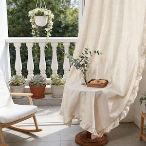 Può includere: Una tenda di lino beige chiaro con un bordo a balze è appesa vicino a un tavolino rotondo coperto da una tovaglia abbinata. Un cestino di vimini con pasticcini è appoggiato sul tavolo. Una sedia a dondolo bianca e piante in vaso sono visibili sul balcone.