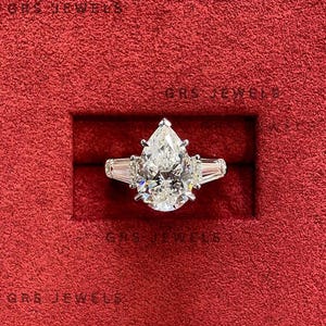 May include: A pear-shaped diamond ring with two baguette-cut side stones, set in a silver band. The ring is displayed in a red jewelry box. The text "GRS JEWELS" is visible on the box.