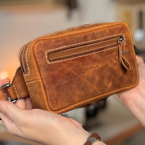 Può includere: Marsupio in pelle marrone con chiusura a zip e cuciture bianche. La borsa rettangolare presenta un piccolo logo. Anche il cinturino è in pelle marrone. Circa 20 cm di larghezza.