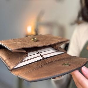 May include: Open brown leather wallet with a snap closure, revealing several white rectangular cards inside. The wallet has a textured, distressed appearance and a dark border. The background is blurred, with a hint of a person holding the wallet.