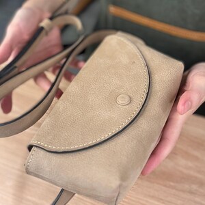 以下が含まれることがあります： ベージュ色のクロスボディバッグ。フラップとボタン留めがあり、表面はテクスチャード加工。長くて調節可能なストラップが付いています。 フラップとバッグ本体の縁にはステッチが施されています。
