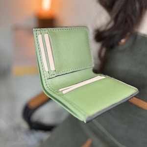 May include: A sage green leather bifold wallet featuring multiple interior card slots and a central compartment. The wallet is constructed with visible dark stitching along the edges and contains several plain white rectangular cards inside.