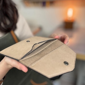 Puede incluir: Cartera de cuero de color beige claro abierta, con múltiples ranuras para tarjetas y un cierre de botón a presión. La cartera se sostiene abierta, mostrando el diseño interior y la construcción. Los bordes están cuidadosamente acabados.