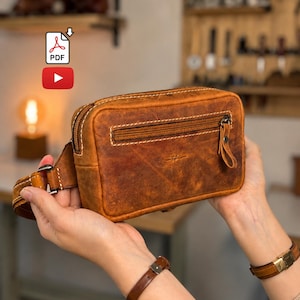 May include: A rectangular brown leather waist bag featuring a distressed finish, a horizontal zippered front pocket with a leather pull tab, a main top zipper, and a matching leather strap with a metal buckle. A small logo is embossed on the front.