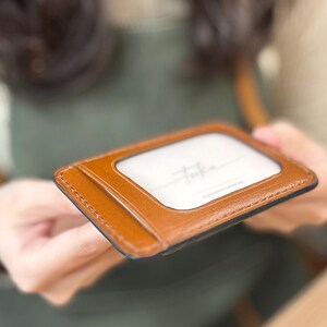 Op de afbeelding: Een bruine leren kaarthouder met een doorzichtig venster, mogelijk voor een ID of creditcards. De kaarthouder heeft een gestikte rand en een zwart interieur. De tekst "Toka" is zichtbaar op de witte kaart binnenin.