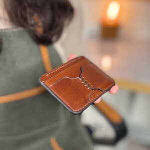 Puede incluir: Un tarjetero de cuero marrón con un diseño cosido. El tarjetero tiene forma rectangular con esquinas redondeadas y múltiples ranuras para tarjetas. El cuero tiene un tono cálido y la costura es visible.
