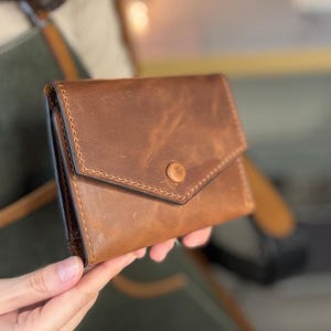 May include: A brown leather wallet with a flap closure and a button clasp. The wallet has a textured surface and visible stitching around the edges. The wallet is held in a person's hand.