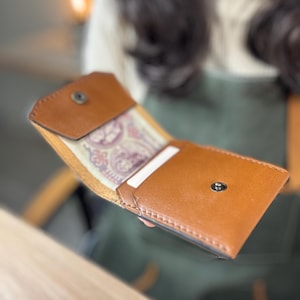 May include: Open, brown leather wallet with a snap closure. The wallet is partially open, revealing a patterned interior and a white card. The leather has a smooth texture and a warm, natural color. The wallet is designed for carrying cards and cash.