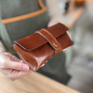 Könnte beinhalten: Eine braune Ledertasche mit Riemenverschluss. Die rechteckige Tasche hat abgerundete Kanten und einen hellbraunen Riemen, der mit einem Knopf befestigt wird. Das Leder hat ein reichhaltiges, strukturiertes Aussehen.