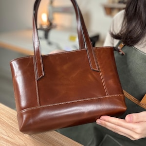 Puede incluir: Un bolso tote de cuero marrón con forma rectangular y dos asas arqueadas. El bolso tiene un acabado liso y brillante y detalles de costura visibles. El bolso es sostenido por una persona.