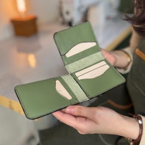 May include: Open olive green leather wallet with multiple card slots and a bill compartment. The wallet is held open, showcasing the interior design and stitching details. The background includes a sewing machine and a warm light.