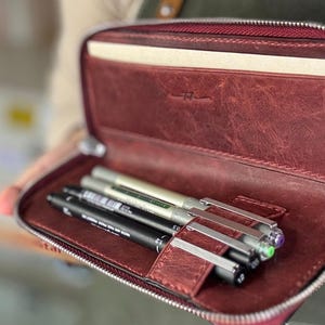 Può includere: Astuccio in pelle bordeaux aperto, con diverse penne nere, argentate e verdi. Le penne sono tenute da un cinturino in pelle. Un taccuino è visibile all'interno dell'astuccio.
