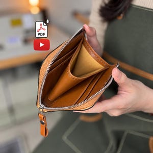 May include: A tan leather wallet with a silver metal zipper closure. The interior features multiple card slots and a central compartment. A leather pull tab is attached to the zipper slider. The wallet is open, revealing the smooth tan lining.