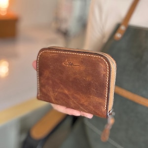 May include: A brown leather wallet with a zip closure. The wallet has a distressed texture and a small logo on the front. The stitching is a light tan colour.