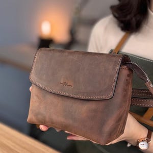 Könnte beinhalten: Braune Leder-Umhängetasche mit Klappenverschluss. Die Tasche hat eine strukturierte Oberfläche und einen genähten Rand. Der Riemen ist seitlich sichtbar. Die Tasche wird von einer Person gehalten.
