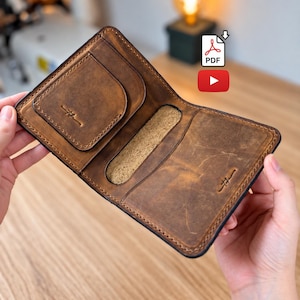 May include: A brown leather bifold wallet featuring visible stitching along the edges, a card slot on the left side, a central vertical opening, and a stamped logo on the right side. The interior displays the natural texture of the leather.