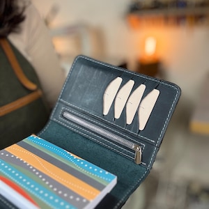 May include: Open leather wallet in dark teal with white stitching and a silver zipper. The wallet holds several light-colored card slots and a small notebook with a colorful striped design.