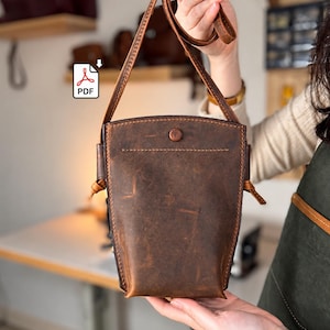 Puede incluir: Un bolso bandolera de cuero marrón oscuro con forma rectangular vertical, que presenta un cierre de botón a presión en la parte superior, costuras visibles en los bordes y una correa larga ajustable con extremos anudados.