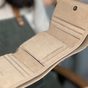 Puede incluir: Cartera bi-fold de color beige claro abierta, con múltiples ranuras para tarjetas y un cierre de botón a presión. La cartera parece estar hecha de cuero o un material similar, con costuras visibles en los bordes. El interior es de un tono beige más claro.