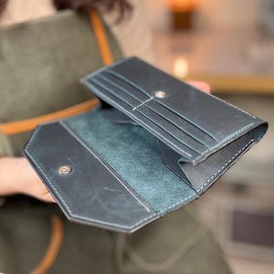 May include: Open, dark blue leather wallet with white stitching and a snap closure. The interior is a lighter blue-green color with card slots. The wallet is open, displaying its contents. The background is blurred.