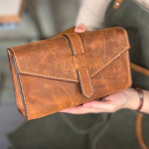 May include: A brown leather clutch with a flap closure and a buckle strap. The clutch has white stitching and a distressed finish. The bag is rectangular and appears to be handmade.