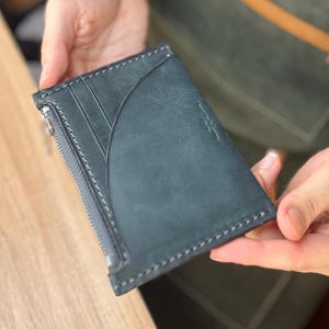 May include: A dark teal leather wallet with a zipper closure and multiple card slots. The wallet has white stitching around the edges and a subtle embossed logo. The wallet is held in a person's hands.