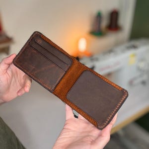 May include: Open brown leather wallet with multiple card slots and a cash compartment. The wallet is held open, showcasing the interior design and stitching details. The leather has a distressed finish.