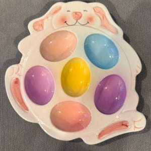 May include: A white ceramic Easter egg holder in the shape of a bunny. The holder has six egg-shaped indentations, each holding a pastel-colored egg: pink, blue, purple, yellow, and orange. The bunny has pink accents on its ears and paws.