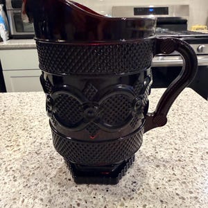 May include: A ruby red glass pitcher with a diamond pattern and decorative accents. The pitcher has a large handle and a spout for easy pouring. The base is square and the pitcher is approximately 9 inches tall.