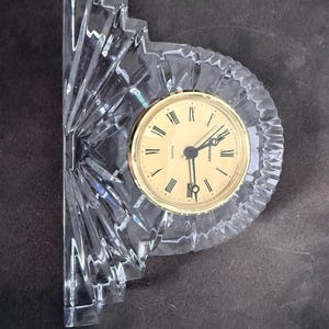Puede incluir: Un reloj de cuarzo de cristal transparente con esfera dorada y manecillas negras. El reloj tiene números romanos y un marco de cristal decorativo en forma de abanico. El reloj mide aproximadamente 15 cm de alto.