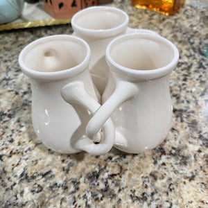 May include: A white ceramic vase with three connected compartments. Each compartment has a rounded top and a handle that intertwines with the others. The vase is set against a speckled countertop.
