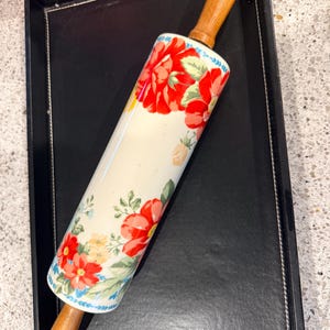 May include: A rolling pin featuring a cylindrical body with a cream-colored background decorated with a vibrant floral pattern of red, yellow, and light blue flowers with green leaves. The rolling pin has smooth, light brown wooden handles.