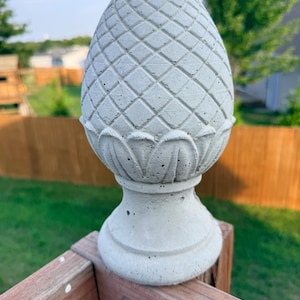 Puede incluir: Un remate de hormigón gris claro con forma de piña, con un patrón de diamantes texturizado en la parte superior y tallas de hojas estilizadas en la base. El objeto presenta una parte superior lisa y redondeada y una base circular.
