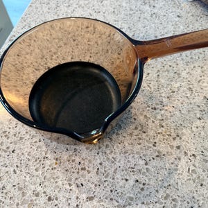 May include: A small, round, transparent amber-coloured glass saucepan featuring a single integrated handle and a small pouring spout on the rim. The interior base of the pan is dark, contrasting with the translucent brown glass walls of the vessel.