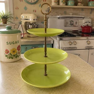 May include: A three-tiered serving stand featuring three round, lime green plates stacked vertically. The plates are connected by a central brass-toned metal rod with a circular handle at the top, designed for displaying food items in a kitchen setting.