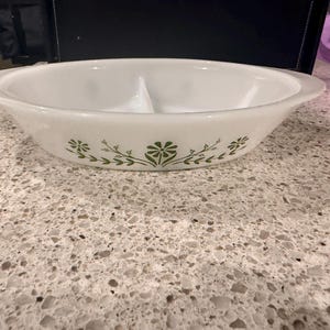 Pode incluir: Uma travessa de servir oval em vidro leitoso branco, dividida em duas partes, com um padrão floral verde no exterior. A peça possui pegas integradas em ambos os lados e um divisor central, criando duas secções separadas para os alimentos.