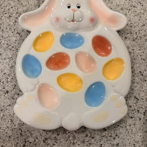 May include: White ceramic Easter bunny-shaped serving platter with colorful egg-shaped compartments. The bunny has pink ears and cheeks, and the egg holders are painted in shades of blue, yellow, orange, and pink. A decorative piece for Easter.